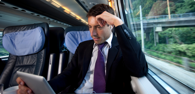 Business man with black suit and purple tie on a train looking down at a silver color tablet.