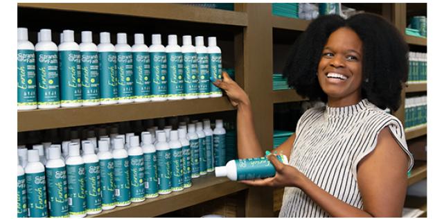 Ameka Coleman, Founder and CEO, is smiling in a black and white-striped shirt. She is standing beside a large shelf full of her leave-in conditioner products.