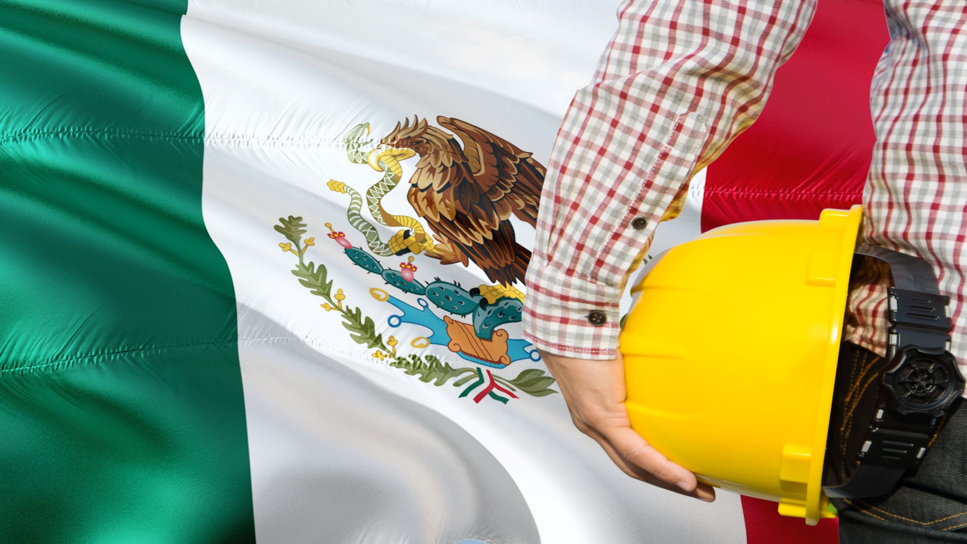 Construction worker with yellow hard hat standing in from of waving Mexico flag.