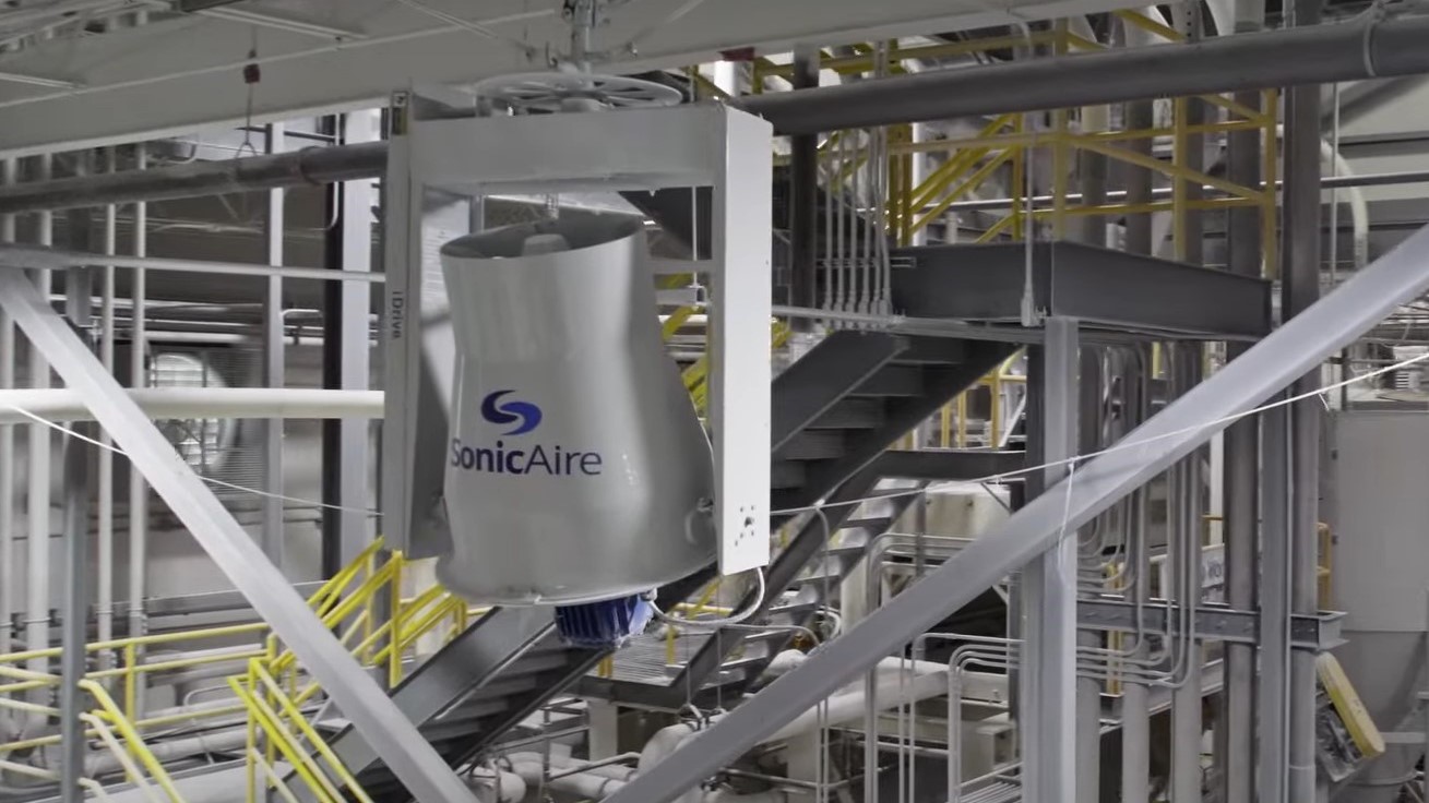 A SonicAire fan is mounted on the ceiling of an empty manufacturing facility that has grey stairs with yellow railings, support beams, and small and medium pipework.