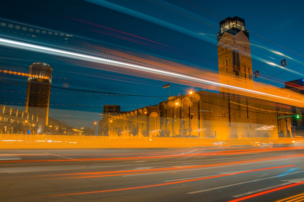 Time lapse photo of traffic at Yerevan city that features blurred lines across a street created from moving vehicles. Featured in the background are some large, historic looking buildings. 