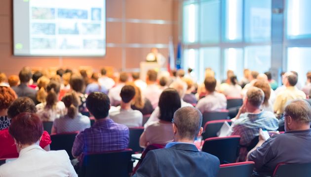 A photograph of a business presenter on a stage presenting to a large audience of business people in business attire.