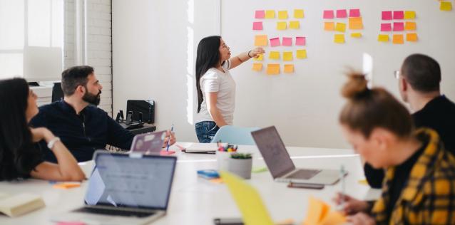 Group of people sitting around table with laptop computers in a meeting room, with one woman standing and pointing at a white board filled with Post-It notes.