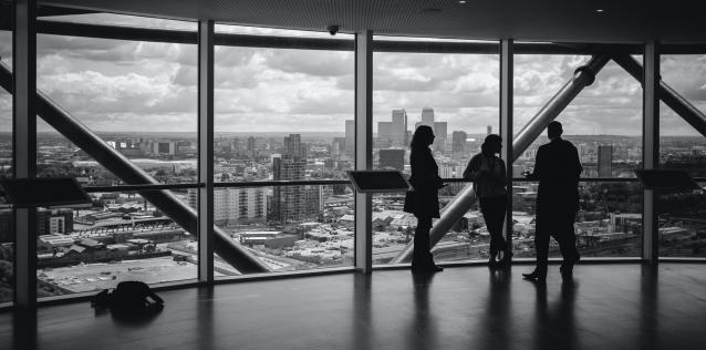There persons having a discussion in front of a large window looking out on a city skyline
