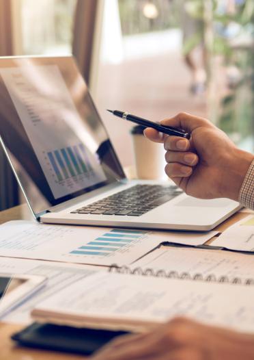 A professionally dressed person holding a pen points at bar charts on laptop screen